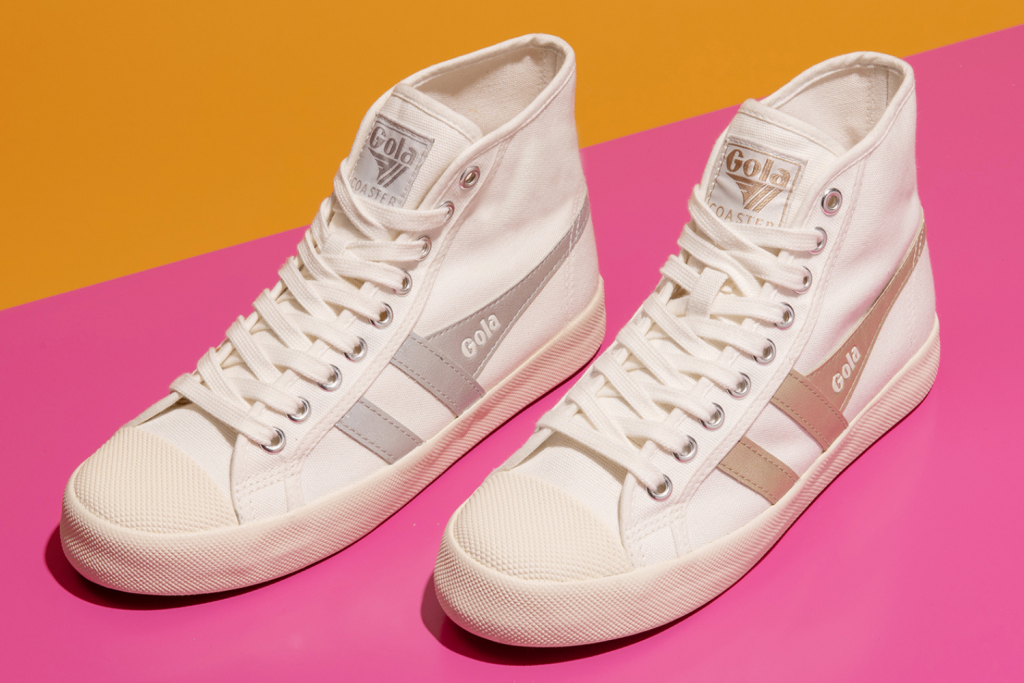Two different styles of Gola high top trainers sit against a contrasting, bold pink and orange striped background. One of the trainers features rose-gold wingflashes, and the other has silver.
