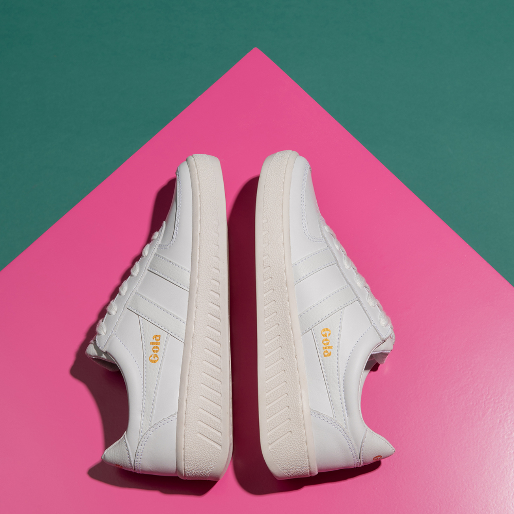 A pair of white trainers are modelled on their side, soles facing eachother. They feature thick white soles with dash detailing, and Gola's logo sits along the wingflash in a chic mustard colour, stylishly contrasting against the white trainer. The background that the trainers are set against is hot pink with dark green and teal detailing in the corners.
